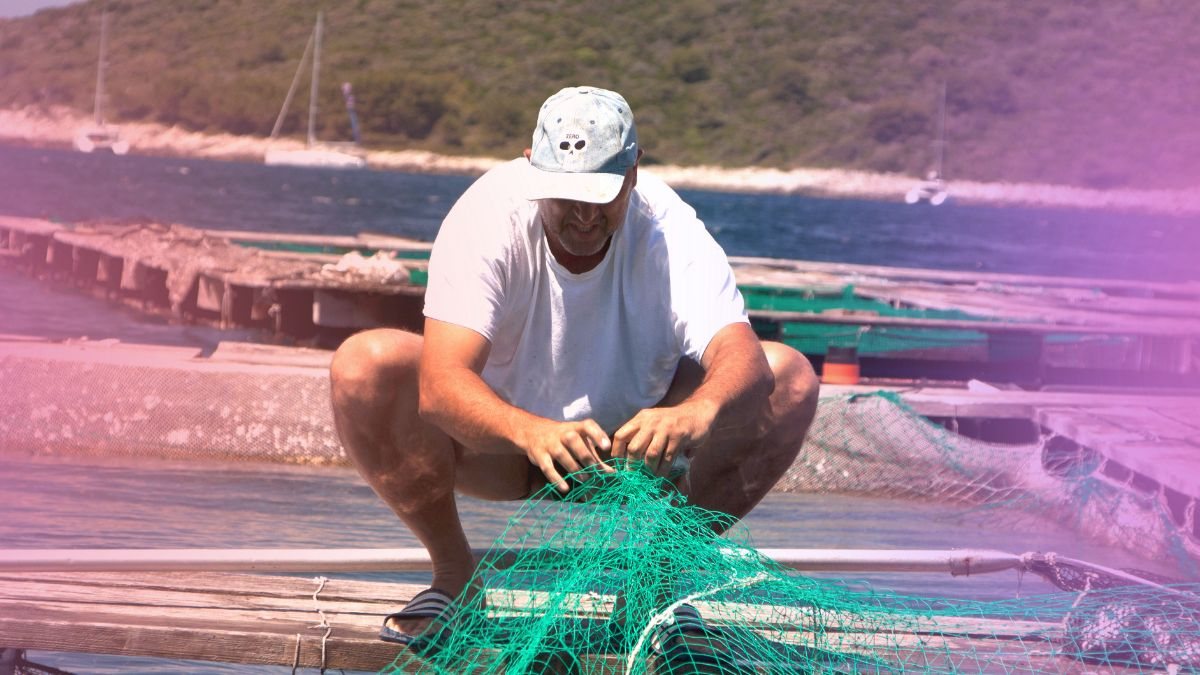 A fisherman mending a net on a wharf with boats moored behind